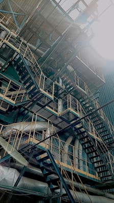 A complex structure of metal staircases, pipes, and railings crisscross within an industrial setting. The sunlight shines directly onto the scene, creating a dramatic contrast and highlighting the intricate network of metalwork.