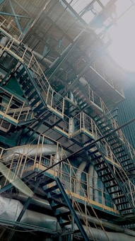 A complex structure of metal staircases, pipes, and railings crisscross within an industrial setting. The sunlight shines directly onto the scene, creating a dramatic contrast and highlighting the intricate network of metalwork.