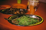 A table set with two oval metal trays of food, one containing bright green vegetables and the other filled with small cooked snails. A drink with a straw, possibly iced tea, is also present in a transparent plastic cup. An orange hue dominates the background with several empty orange plates visible.