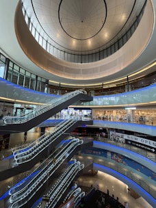 A multi-level shopping mall interior with several escalators leading to different floors. The architecture features a large, circular ceiling with ample glass windows allowing natural light in. Various shops with illuminated signs are visible on the upper levels.