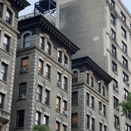 Multiple stories of old, classic style apartment buildings with intricate architectural details. The facades feature large windows and air conditioning units. The brick buildings are a mix of light and dark tones with some greenery visible at the bottom.