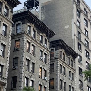 Multiple stories of old, classic style apartment buildings with intricate architectural details. The facades feature large windows and air conditioning units. The brick buildings are a mix of light and dark tones with some greenery visible at the bottom.