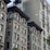 Multiple stories of old, classic style apartment buildings with intricate architectural details. The facades feature large windows and air conditioning units. The brick buildings are a mix of light and dark tones with some greenery visible at the bottom.