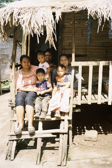 A family is sitting on the steps of a traditional wooden house with a thatched roof. There are two adults and four children, all appearing relaxed and content. The house and surroundings have a rustic, rural feel with natural materials and simple construction.