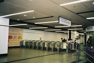 An indoor setting with a row of turnstiles positioned in front of a group of people. The ceiling features linear lighting strips, and there are signs in the area indicating directions. A promotional poster for a loyalty programme is visible on the wall.