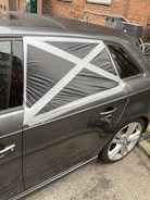 A car with a damaged rear window partially covered by a makeshift repair using black plastic and silver duct tape, arranged in a cross pattern. The vehicle is parked on a street with a brick wall and some utility items in the background.