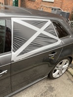 A car with a damaged rear window partially covered by a makeshift repair using black plastic and silver duct tape, arranged in a cross pattern. The vehicle is parked on a street with a brick wall and some utility items in the background.