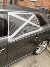 A car with a damaged rear window partially covered by a makeshift repair using black plastic and silver duct tape, arranged in a cross pattern. The vehicle is parked on a street with a brick wall and some utility items in the background.