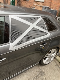 A car with a damaged rear window partially covered by a makeshift repair using black plastic and silver duct tape, arranged in a cross pattern. The vehicle is parked on a street with a brick wall and some utility items in the background.