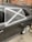 A car with a damaged rear window partially covered by a makeshift repair using black plastic and silver duct tape, arranged in a cross pattern. The vehicle is parked on a street with a brick wall and some utility items in the background.