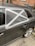 A car with a damaged rear window partially covered by a makeshift repair using black plastic and silver duct tape, arranged in a cross pattern. The vehicle is parked on a street with a brick wall and some utility items in the background.