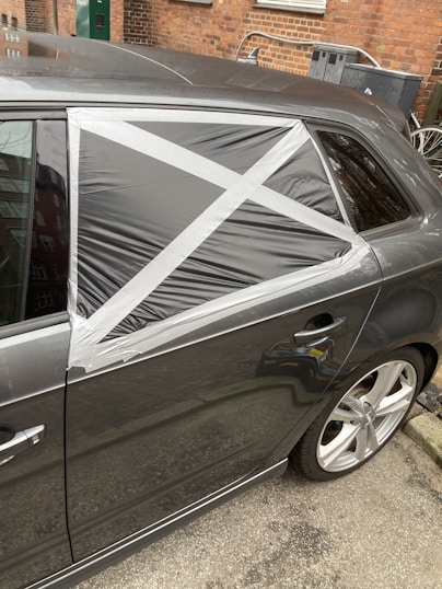 A car with a damaged rear window partially covered by a makeshift repair using black plastic and silver duct tape, arranged in a cross pattern. The vehicle is parked on a street with a brick wall and some utility items in the background.