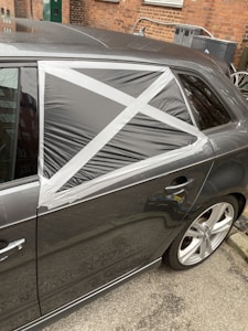 A car with a damaged rear window partially covered by a makeshift repair using black plastic and silver duct tape, arranged in a cross pattern. The vehicle is parked on a street with a brick wall and some utility items in the background.
