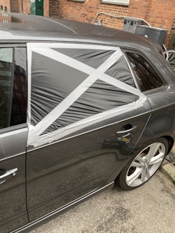 A car with a damaged rear window partially covered by a makeshift repair using black plastic and silver duct tape, arranged in a cross pattern. The vehicle is parked on a street with a brick wall and some utility items in the background.