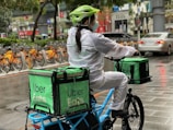A person wearing a helmet and a mask rides a bicycle, carrying large green delivery bags labeled 'Uber Eats.' The surroundings show a row of parked orange bicycles, a wet pavement, and various urban elements like cars and street signs.