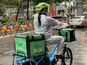 A person wearing a helmet and a mask rides a bicycle, carrying large green delivery bags labeled 'Uber Eats.' The surroundings show a row of parked orange bicycles, a wet pavement, and various urban elements like cars and street signs.