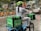 A person wearing a helmet and a mask rides a bicycle, carrying large green delivery bags labeled 'Uber Eats.' The surroundings show a row of parked orange bicycles, a wet pavement, and various urban elements like cars and street signs.
