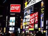 A vibrant urban scene featuring multiple illuminated advertisements in a cityscape. Brightly lit billboards display various brand names and logos, including Coca-Cola, against a nighttime backdrop. Neon signs and text in different languages add to the bustling atmosphere. The architecture and presence of diverse signs suggest a busy commercial district.