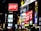 A vibrant urban scene featuring multiple illuminated advertisements in a cityscape. Brightly lit billboards display various brand names and logos, including Coca-Cola, against a nighttime backdrop. Neon signs and text in different languages add to the bustling atmosphere. The architecture and presence of diverse signs suggest a busy commercial district.