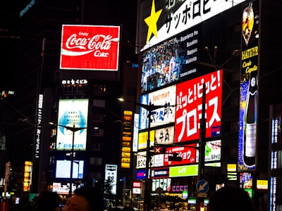 A vibrant urban scene featuring multiple illuminated advertisements in a cityscape. Brightly lit billboards display various brand names and logos, including Coca-Cola, against a nighttime backdrop. Neon signs and text in different languages add to the bustling atmosphere. The architecture and presence of diverse signs suggest a busy commercial district.