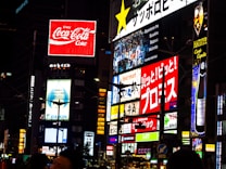 A vibrant urban scene featuring multiple illuminated advertisements in a cityscape. Brightly lit billboards display various brand names and logos, including Coca-Cola, against a nighttime backdrop. Neon signs and text in different languages add to the bustling atmosphere. The architecture and presence of diverse signs suggest a busy commercial district.