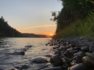 A serene riverside scene at sunset, with smooth pebbles in the foreground and still water reflecting the golden hues of the setting sun. Trees line the riverbank, casting dark silhouettes against the colorful sky.