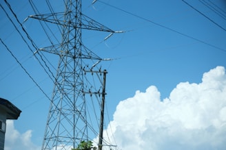 A tall metal transmission tower stands against a bright blue sky with fluffy white clouds. Several power lines extend from the tower, intersecting and leading off-frame. A part of a building roof is visible in the lower left corner with some greenery below the tower.