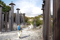 A person with a blue backpack is walking through an area adorned with tall wooden pillars inscribed with Japanese characters. The surroundings are natural, with greenery and a gravel path leading through the structures. In the distance, a traditional stone monument is visible against a backdrop of hills and a partly cloudy sky.