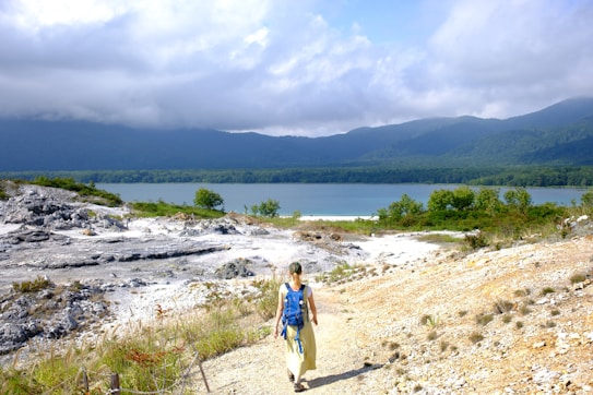 A person with a backpack walks along a rocky path towards a tranquil lakeside landscape, with vibrant green foliage and a backdrop of mountains partially shrouded by clouds.