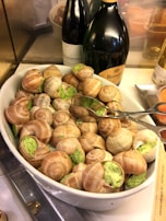 A large white bowl contains escargots filled with a green herb butter mixture. The shells are arranged neatly, displaying the appetizing herb filling. Behind the bowl, there are bottles of wine or champagne, including a prominent dark bottle with the label 'Ruinart'. A pair of metal tongs rests on top of the escargots, suggesting the dish is ready to be served.
