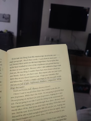 An open book held in a room, with text highlighting themes of self-worth and inner beauty. The background includes a television mounted on a wall, a piece of furniture, and a partially visible doorway, suggesting a cozy, indoor setting.