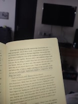 An open book held in a room, with text highlighting themes of self-worth and inner beauty. The background includes a television mounted on a wall, a piece of furniture, and a partially visible doorway, suggesting a cozy, indoor setting.