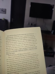 An open book held in a room, with text highlighting themes of self-worth and inner beauty. The background includes a television mounted on a wall, a piece of furniture, and a partially visible doorway, suggesting a cozy, indoor setting.