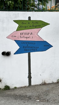 A signpost is painted on a white wall with three colorful directional arrows. The top green arrow points to the left and reads 'Casa da Árvore Albergue dos Peregrinos 5m'. The middle pink arrow points to the right and reads 'Fátima (Portugal) 171 km'. The bottom blue arrow points to the left and reads 'Santiago de Compostela (España) 262 km'. There are plants and flowers visible above the wall and some greenery and pipes at the bottom left.