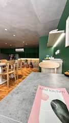 A modern kitchen setting with a mix of green and natural wood tones. There's a dining table with wooden chairs, several potted plants, and an air conditioning unit on the wall. A countertop displays various items including a bottle. The ceiling has a textured gray finish, and a pink and green flyer or magazine titled 'kind kitchen' lies on the foreground surface.