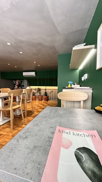 A modern kitchen setting with a mix of green and natural wood tones. There's a dining table with wooden chairs, several potted plants, and an air conditioning unit on the wall. A countertop displays various items including a bottle. The ceiling has a textured gray finish, and a pink and green flyer or magazine titled 'kind kitchen' lies on the foreground surface.