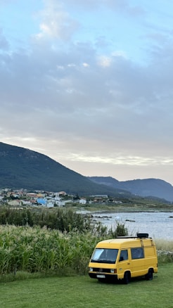 A yellow van is parked on a grassy field near a body of water. In the background, there is a town with colorful houses nestled at the base of green hills. The sky is cloudy, creating a serene and calm atmosphere.
