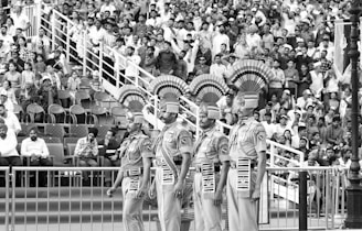 A uniformed parade with four soldiers standing in a line, displaying ornate headgear, in front of a large seated crowd. The soldiers wear decorated ceremonial outfits. The crowd is engaged and watching attentively.