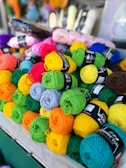 A collection of multi-colored yarn balls neatly stacked on a table. The yarn balls come in various colors including red, green, blue, yellow, pink, orange, and gray. They are arranged in a visually appealing manner, with labels intact, suggesting they are possibly for sale. The background shows additional yarns and a slightly blurred setting, likely a store or market.