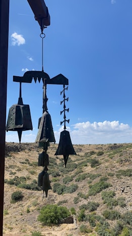 Metal wind chimes hang against a backdrop of a wide, open landscape. The chimes are intricately designed with various shapes, suspended from a wooden beam. The landscape features rolling hills covered in sparse shrubbery under a bright blue sky with scattered clouds.
