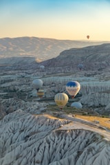 Hot air balloons float above a scenic landscape with unique rock formations and valleys, capturing the beauty of a sunrise or sunset. The balloons are colorful and diverse, some with inscriptions, and are surrounded by vast mountains and a clear sky.