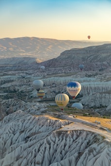 Hot air balloons float above a scenic landscape with unique rock formations and valleys, capturing the beauty of a sunrise or sunset. The balloons are colorful and diverse, some with inscriptions, and are surrounded by vast mountains and a clear sky.