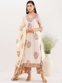 A woman is wearing a traditional ethnic outfit with floral patterns. The ensemble includes a long tunic with matching pants and a draped scarf. The color scheme is primarily cream with pink and green floral designs. She is standing against a plain background with a large white vase containing green foliage beside her. Her hair is straight and she is wearing large silver earrings.