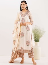 A woman is wearing a traditional ethnic outfit with floral patterns. The ensemble includes a long tunic with matching pants and a draped scarf. The color scheme is primarily cream with pink and green floral designs. She is standing against a plain background with a large white vase containing green foliage beside her. Her hair is straight and she is wearing large silver earrings.