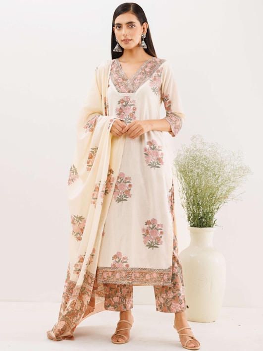 A woman is wearing a traditional ethnic outfit with floral patterns. The ensemble includes a long tunic with matching pants and a draped scarf. The color scheme is primarily cream with pink and green floral designs. She is standing against a plain background with a large white vase containing green foliage beside her. Her hair is straight and she is wearing large silver earrings.