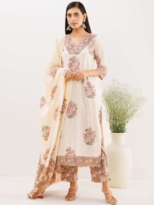 A woman is wearing a traditional ethnic outfit with floral patterns. The ensemble includes a long tunic with matching pants and a draped scarf. The color scheme is primarily cream with pink and green floral designs. She is standing against a plain background with a large white vase containing green foliage beside her. Her hair is straight and she is wearing large silver earrings.