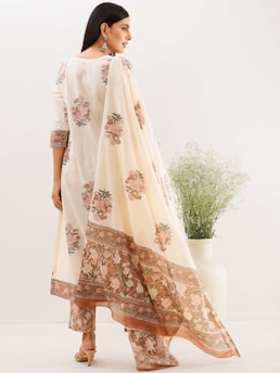 A woman is standing in a light-colored traditional outfit with floral patterns. The attire includes a flowy dupatta draped over her shoulder. She is adorned with large earrings and wearing open-toe heels. In the background, there is a large green plant in a white pot, and the setting has a minimalistic aesthetic.