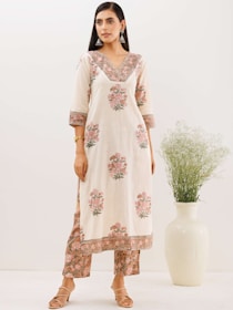 A woman is standing in a light-colored, floral patterned kurta and pants set, featuring intricate designs with pink and green hues. She has long straight hair and is wearing large silver earrings and a bracelet. She stands next to a tall white vase with green foliage.