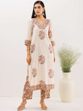 A woman is standing in a light-colored, floral patterned kurta and pants set, featuring intricate designs with pink and green hues. She has long straight hair and is wearing large silver earrings and a bracelet. She stands next to a tall white vase with green foliage.