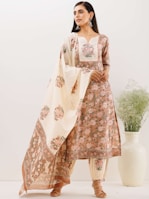 A woman stands wearing a traditional floral embroidered outfit consisting of a kurta, pants, and a dupatta. The ensemble features intricate patterns and soft, pastel colors. She accessorizes with large earrings and sandals, standing beside a white decorative vase with green foliage.